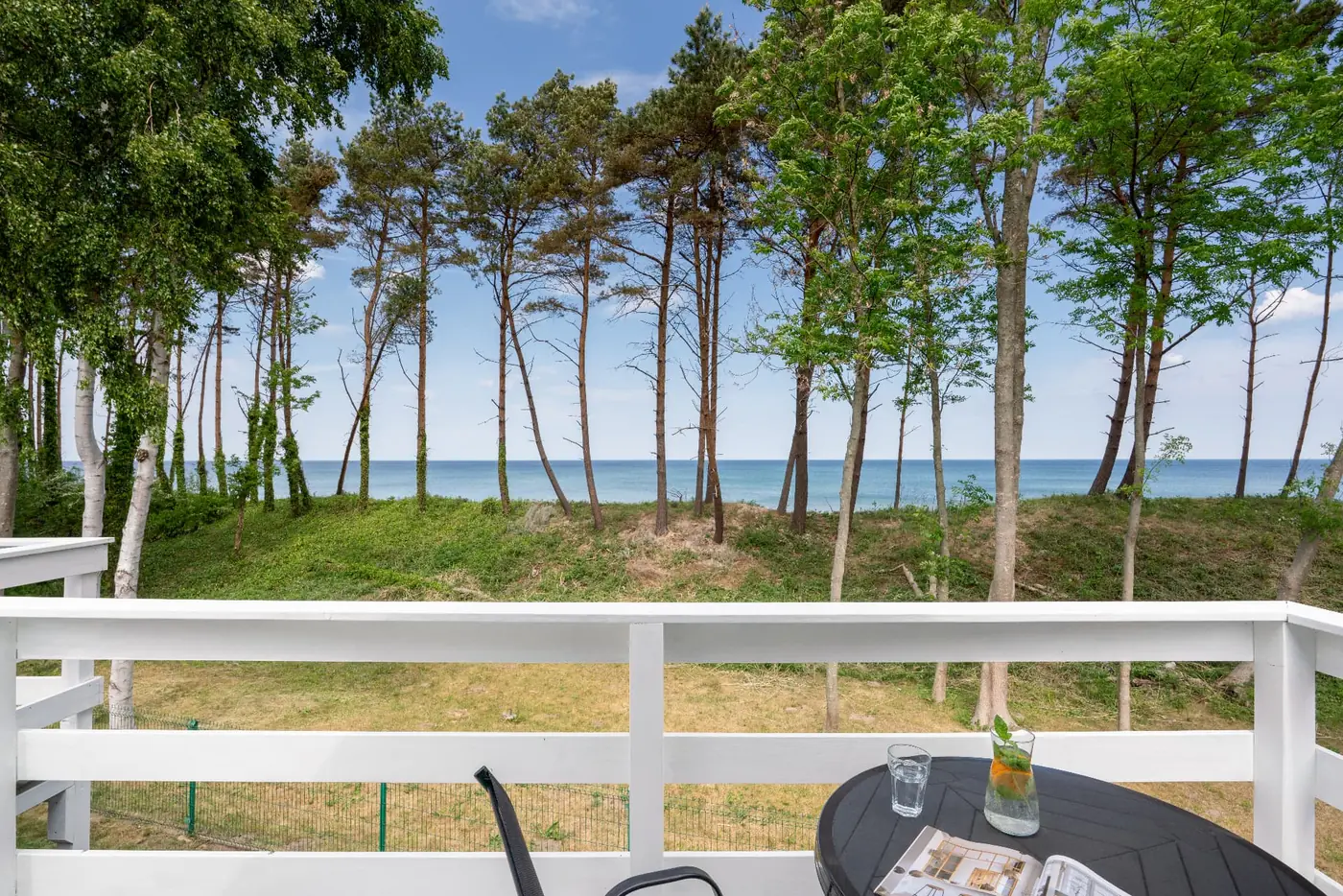 Balkon z widokiem na morze – domki Sarbinowo Balkon domku letniskowego w Sarbinowie ze stolikiem i widokiem na morze przez sosny