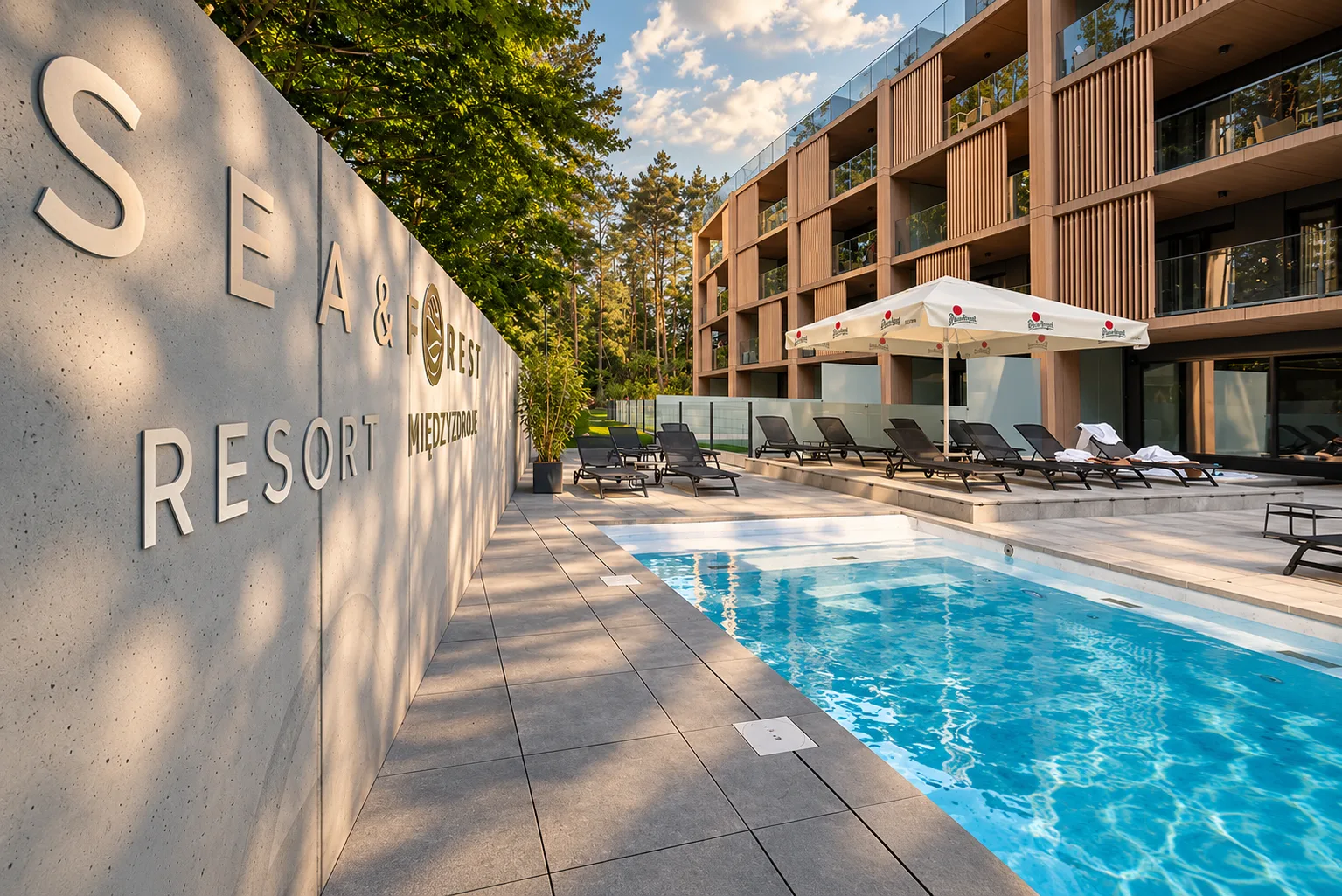 Betonowa ściana z logo przy basenie zewnętrznym i budynkiem resortu Sea & Forest